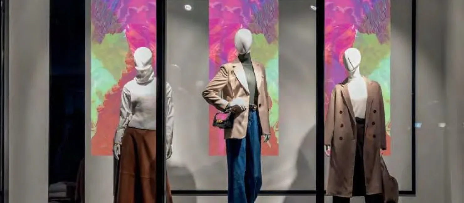 Fashion storefront at night: three mannequins stand in front of tall dichroic panels glowing pink and green behind the glass
